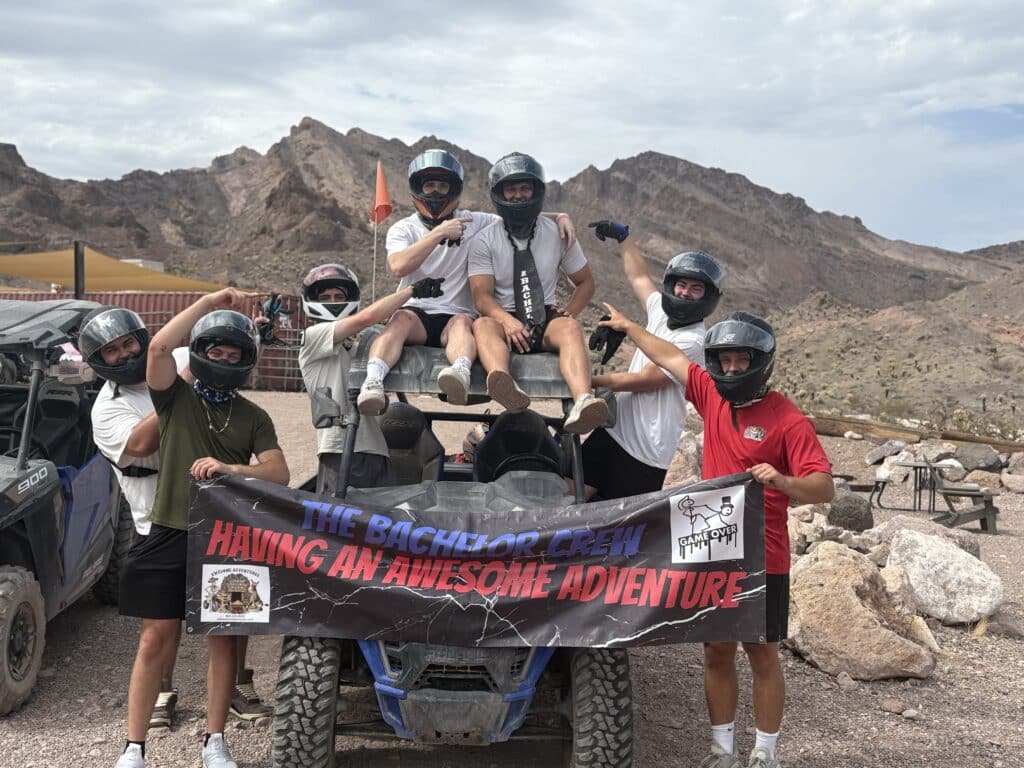 Group of men in helmets pose by ATV in Nevada desert near Las Vegas, holding Bachelor Crew banner during RZR off-road adventure.