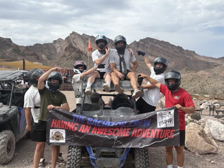 Six men in helmets pose with a banner on an ATV tour near Las Vegas in the Nevada desert, mountains and clouds behind.