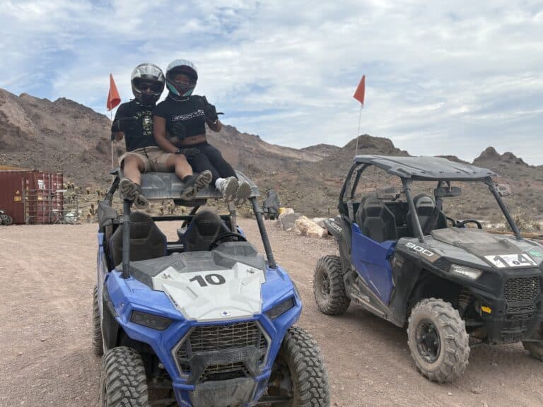 Riders wearing helmets on a blue ATV #10 in Nevada desert near Las Vegas; ATV tours, rocky terrain, distant mountains visible.