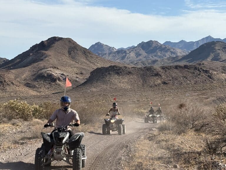 ATV riders explore a dusty Nevada desert trail near Las Vegas with red flags, rocky hills, and mountains in the background.