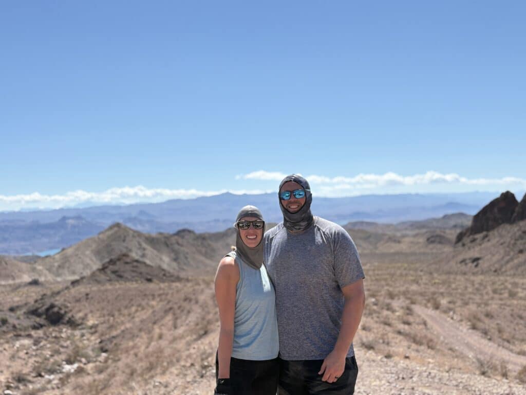 Two people in sun hoods and sunglasses pose in a Nevada desert near Las Vegas, with a lake and mountains—ideal for ATV tours.