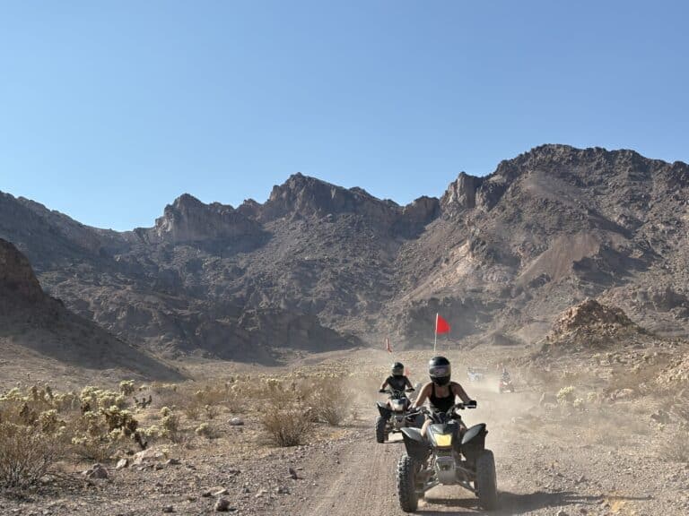ATV tour riders kick up dust on a Nevada desert trail near Las Vegas, red flags flying, with mountains and blue sky in the background.