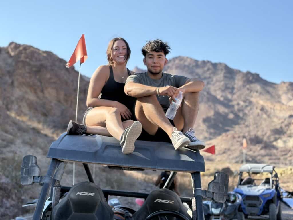 Smiling friends atop an ATV near Nevada’s rocky mountains on a sunny RZR off-road tour; Las Vegas adventure in background.