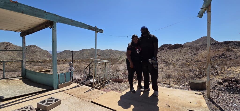 Two people on a wooden platform by a blue-framed structure near Las Vegas, Nevada, with desert and mountain views—ideal for ghost town tours.