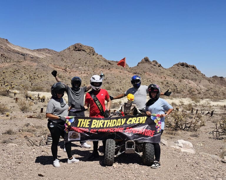 Group in helmets holds THE BIRTHDAY CREW banner by ATV in Nevada desert near Las Vegas, ready for an epic RZR off-road adventure.
