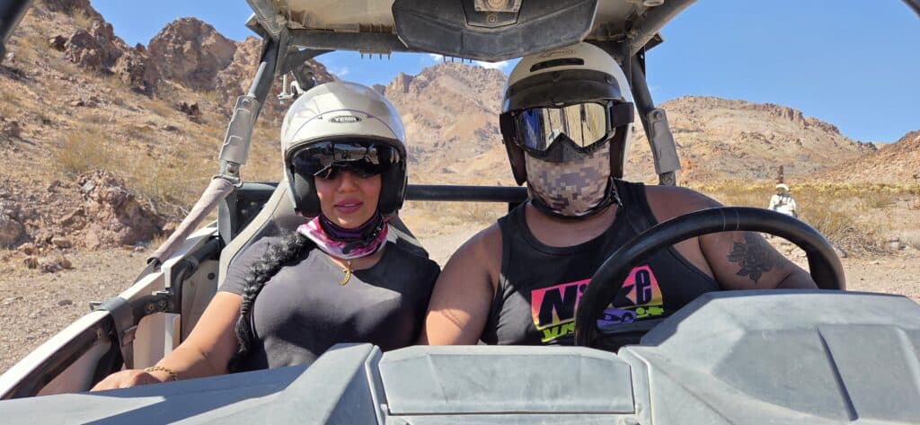 Two people on a Nevada ATV tour ride an RZR in the desert near Las Vegas; rocky hills, blue sky, and pixelated face visible.
