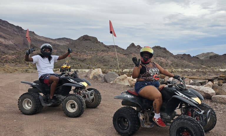 Adventurers on ATVs flash peace signs during a Nevada desert ATV tour near Las Vegas, rocky trails and mountains behind them.