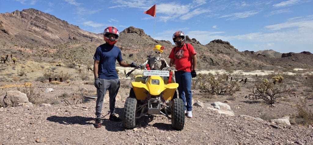 Las Vegas ATV tour: Two riders and a skeleton on a yellow ATV in the Nevada desert, red flag waving, mountains in the background.