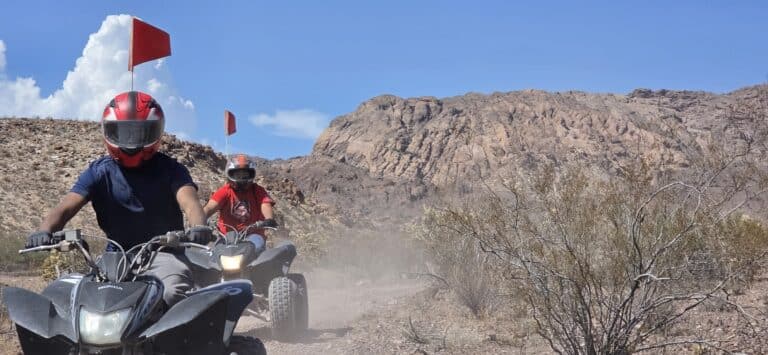 ATV riders speed along a dusty Nevada desert trail near Las Vegas, with red safety flags and rocky mountains in the background.