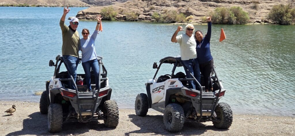 Group poses with raised arms by RZRs near Colorado River, Las Vegas; Nevada hills, shrubs, and a duck complete this ATV tour scene.