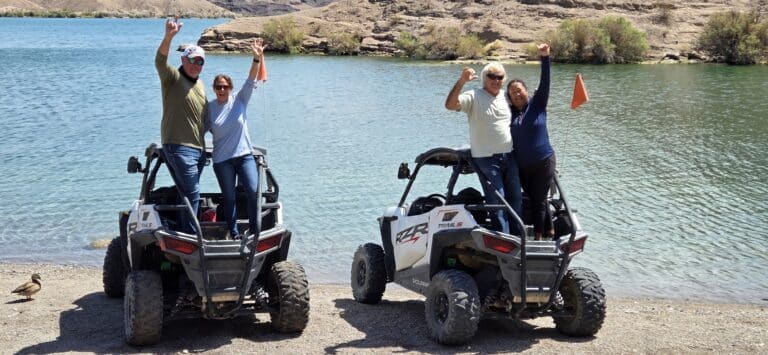 Group poses with raised arms by RZRs near Colorado River, Las Vegas; Nevada hills, shrubs, and a duck complete this ATV tour scene.