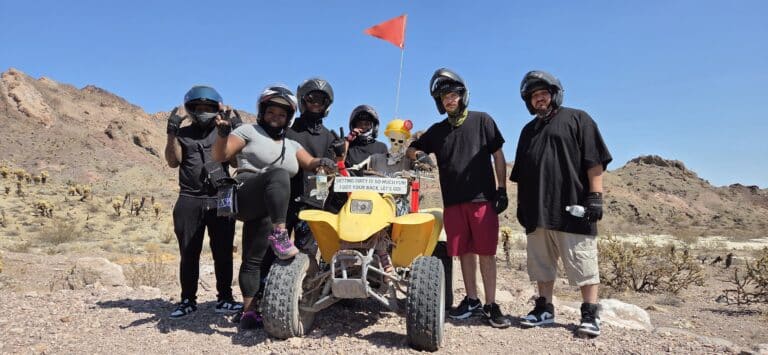 Six adventurers pose by a yellow ATV with red flag on a Nevada desert ATV tour near Las Vegas, rocky hills and blue sky behind.