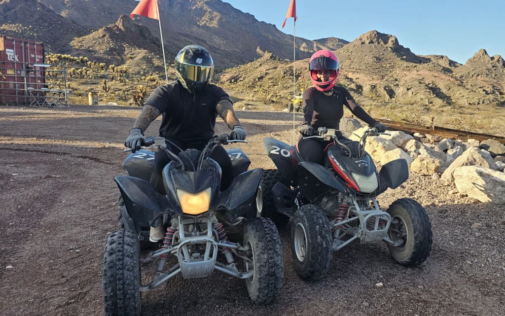 Riders in black gear on ATVs rest in Nevada desert near Las Vegas, with mountains and red flags—perfect ATV tour adventure.