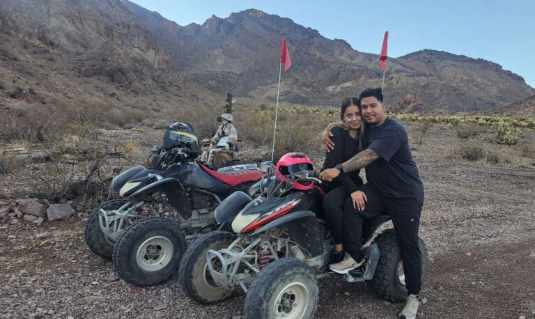 Smiling couple on ATV during a Nevada desert RZR tour near Las Vegas, with mountains and extra ATVs in the background.