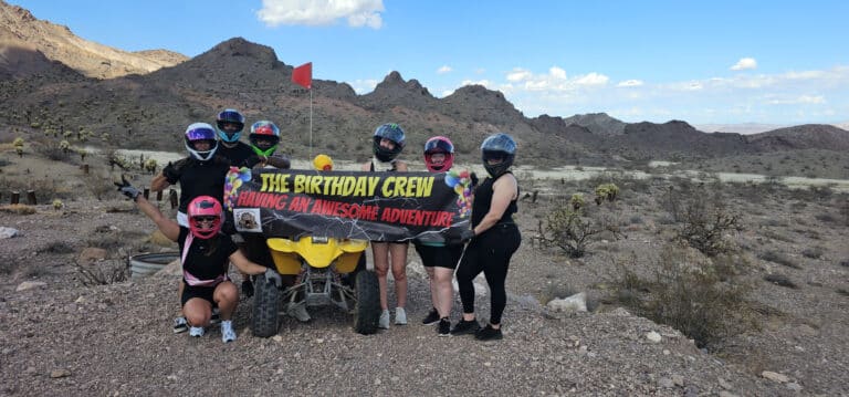 Six friends in helmets celebrate with a banner by a yellow ATV on a Nevada desert tour near Las Vegas mountains.
