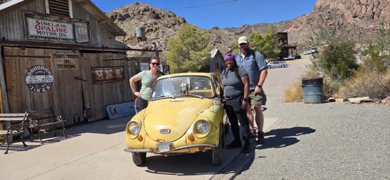 Three people by a vintage yellow car at a Nevada ghost town near Las Vegas, rustic signs, desert hills—perfect for ATV tours.