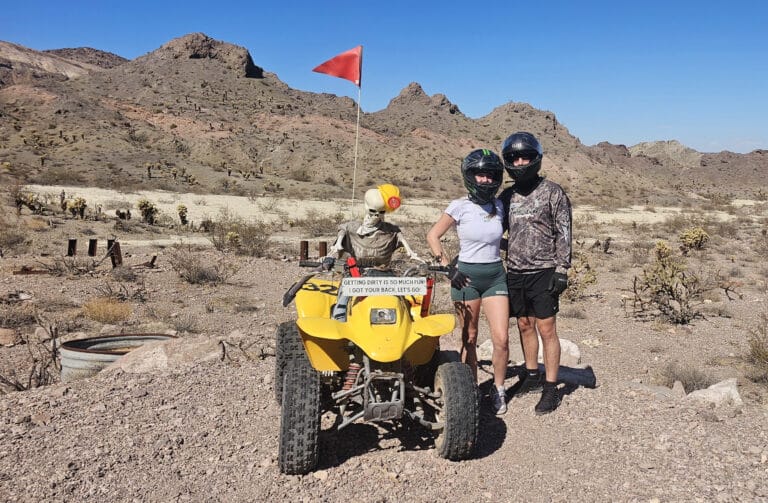 ATV tour near Las Vegas: two riders in helmets beside a yellow quad with skeleton at the wheel, Nevada desert mountains behind.