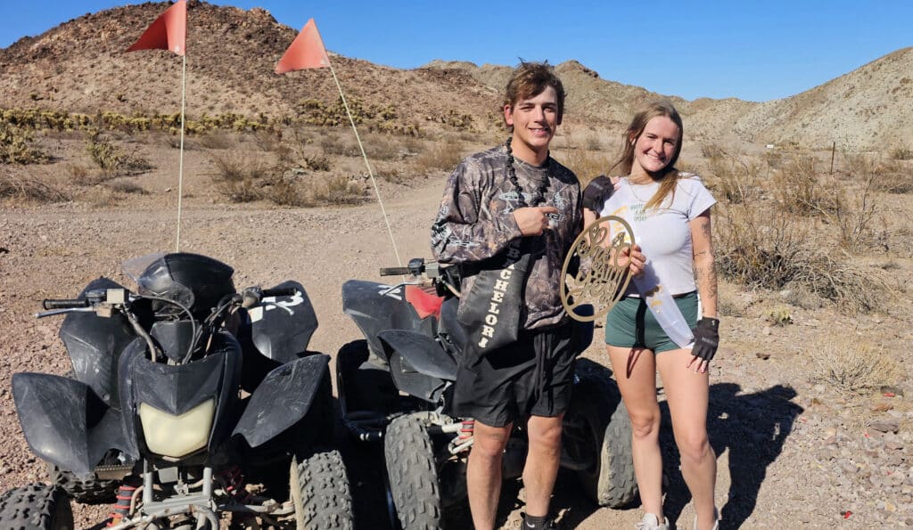 Two people smile by ATVs in a Nevada desert near Las Vegas, ready for an ATV tour with hills and adventure gear visible.