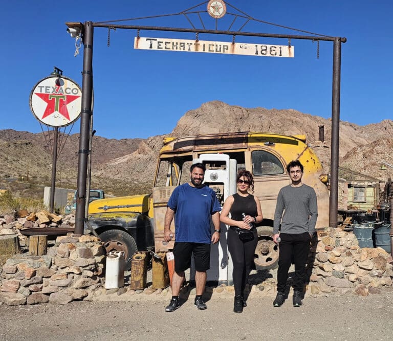 Three people by a rusty yellow truck at Techatticup Ghost Town near Las Vegas, Nevada. Rocky hills and old signs set the scene.