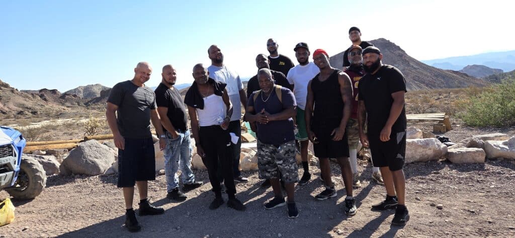 Thirteen men pose on rocky Nevada terrain near Las Vegas, with mountains behind, dressed for an ATV tour under a clear blue sky.