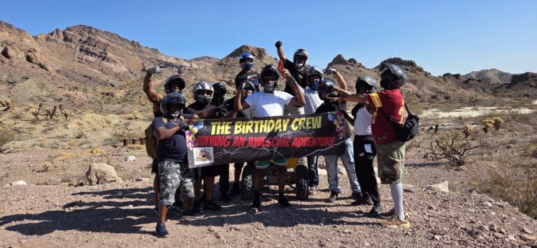 Adventure group in helmets poses in Nevada desert by rocky hills, holding "THE BIRTHDAY CREW" banner after RZR off-road tour near Las Vegas.