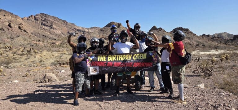 Smiling group in helmets and masks raises fists on a Nevada desert ATV tour, holding THE BIRTHDAY CREW banner near Las Vegas.