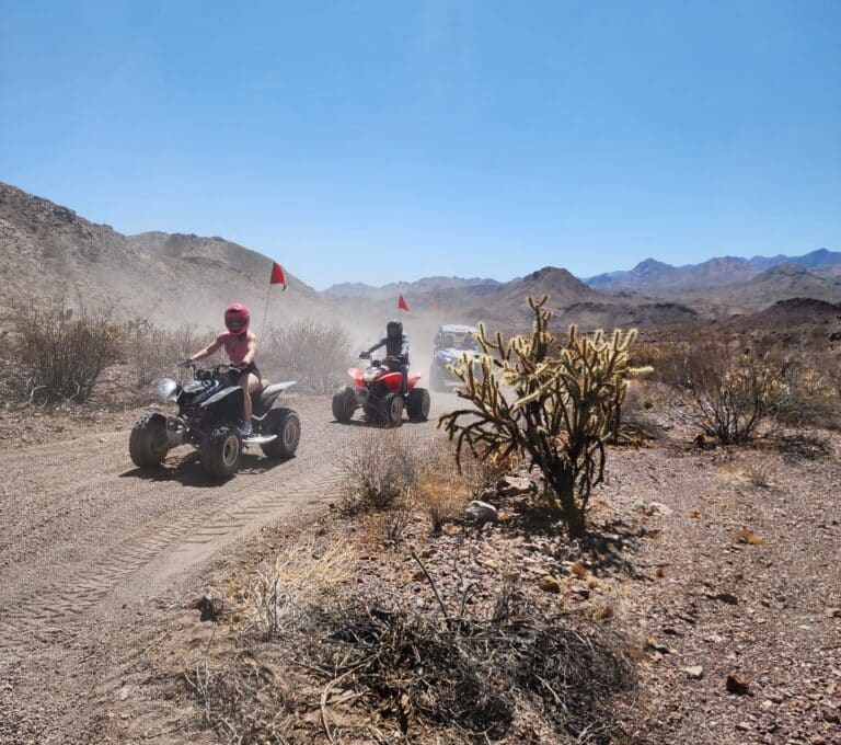Riders enjoy an ATV tour on a dusty Nevada desert trail near Las Vegas, red flags flying for safety with mountains in the background.