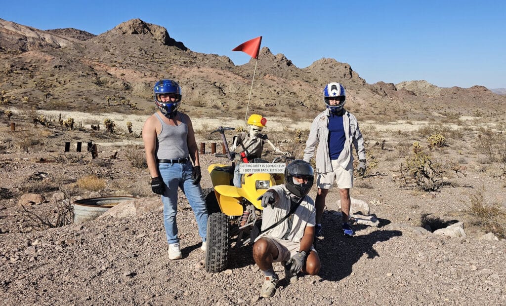 Three people in helmets pose by a yellow ATV on a Nevada desert tour near Las Vegas, with mountains and rocky terrain behind them.
