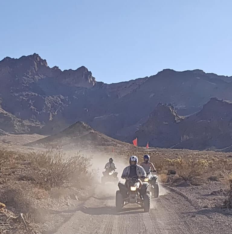ATV tour near Las Vegas: three riders with red flags drive desert trails, mountains and blue sky in Nevada backdrop.