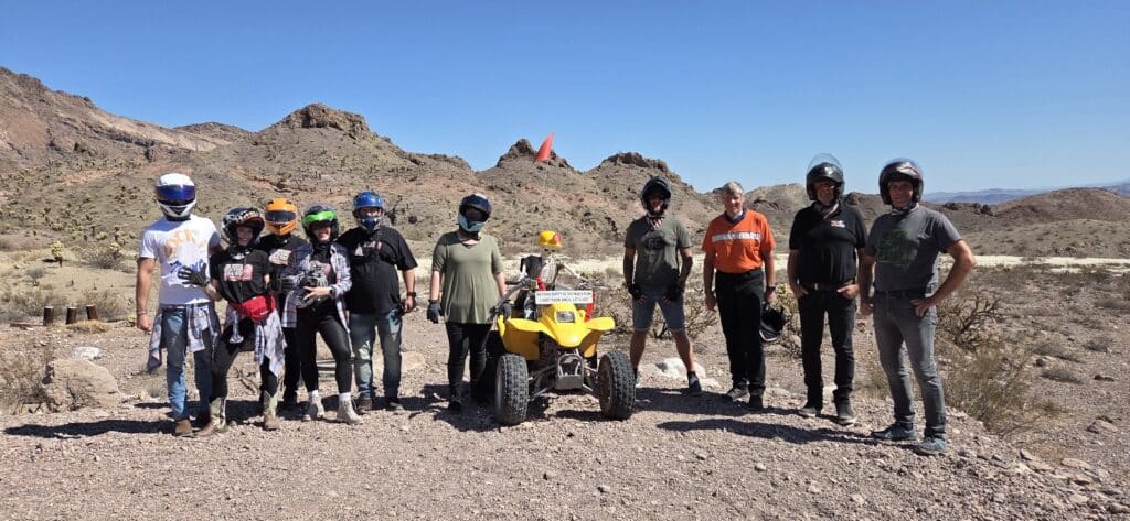 Nine riders in helmets on Nevada desert terrain by a yellow ATV, near Las Vegas, ready for an ATV tour with scenic mountain views.