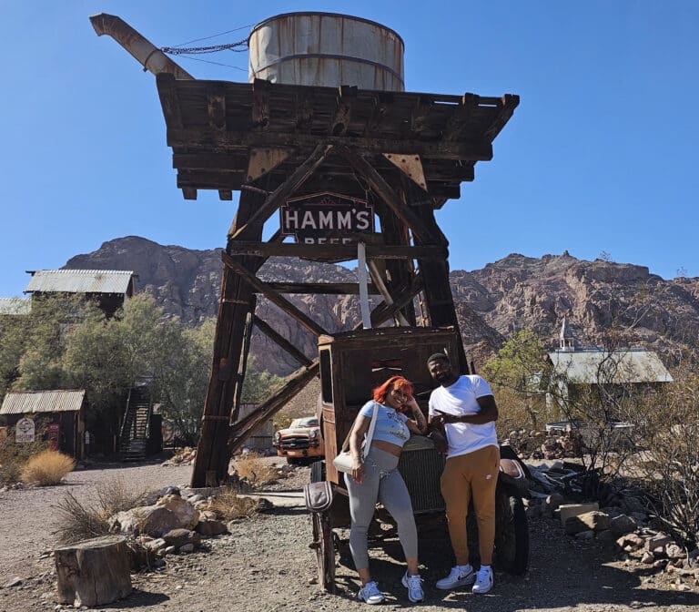 Smiling visitors pose by a HAMMS BEER water tower in a Nevada ghost town near the Colorado River and Las Vegas desert scenery.