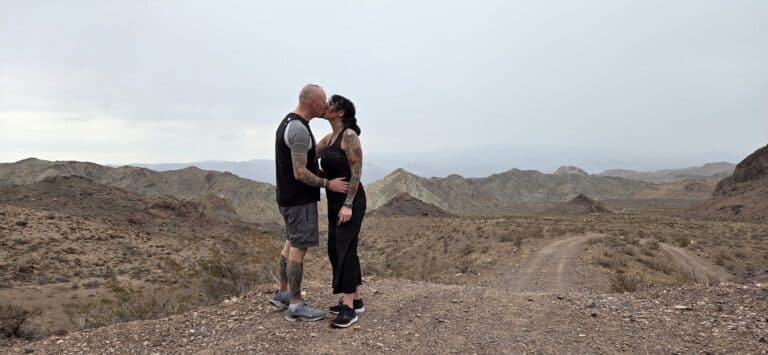 Tattooed couple kisses on a rocky Nevada desert hill near Las Vegas, with dry mountains and overcast sky in the background.