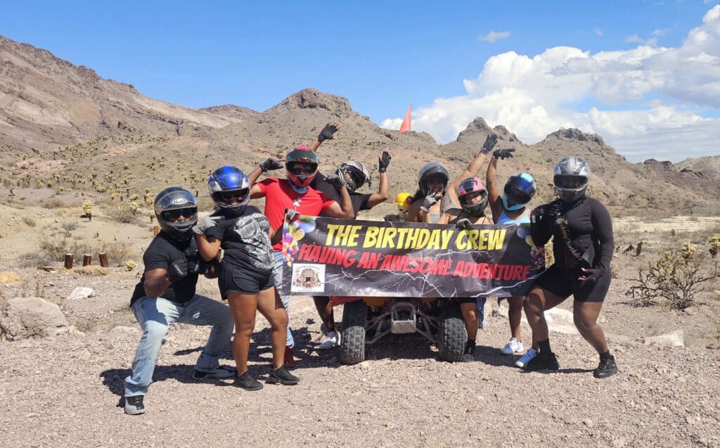 Seven friends in helmets pose by ATVs, holding banner on a Las Vegas ATV tour near Nevada’s rocky desert and Colorado River.