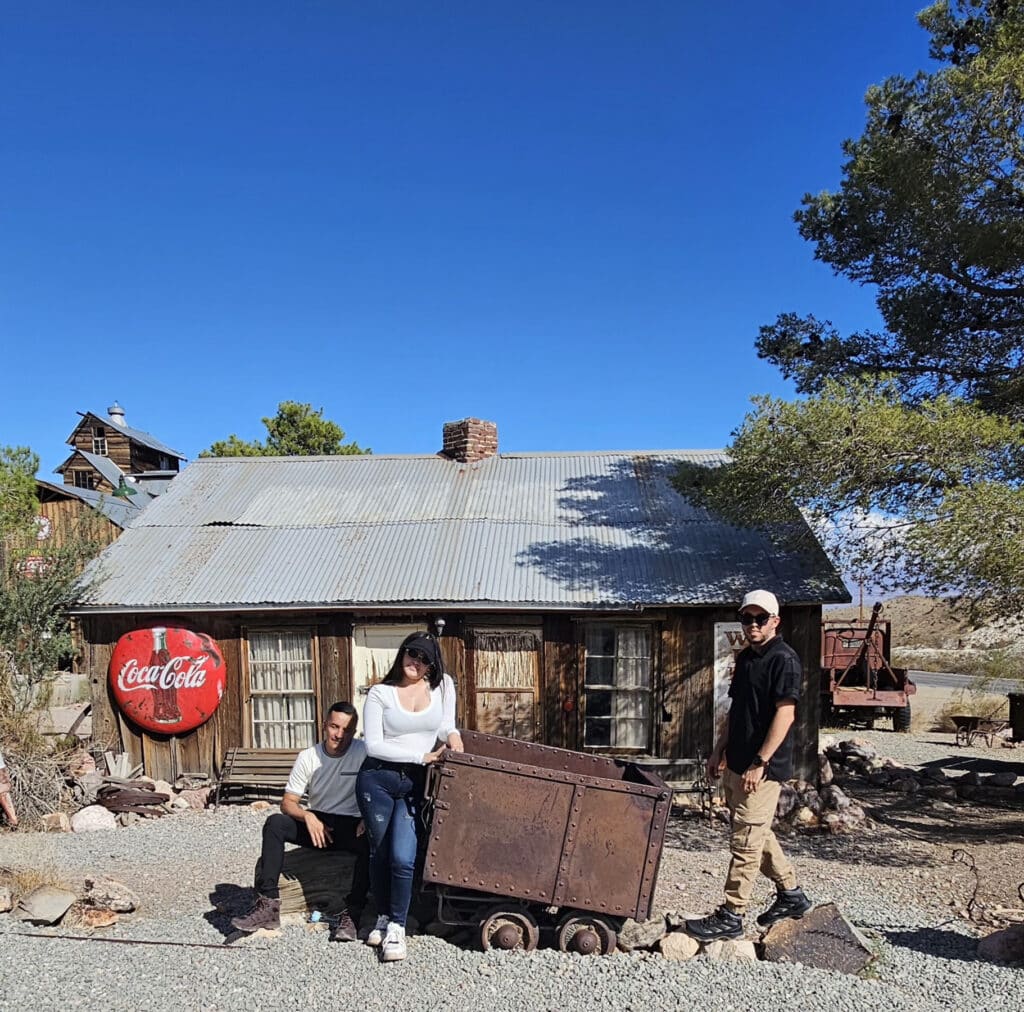 Three people pose by a mining cart at a Nevada ghost town near Las Vegas, with vintage signs—perfect for sightseeing and photos.