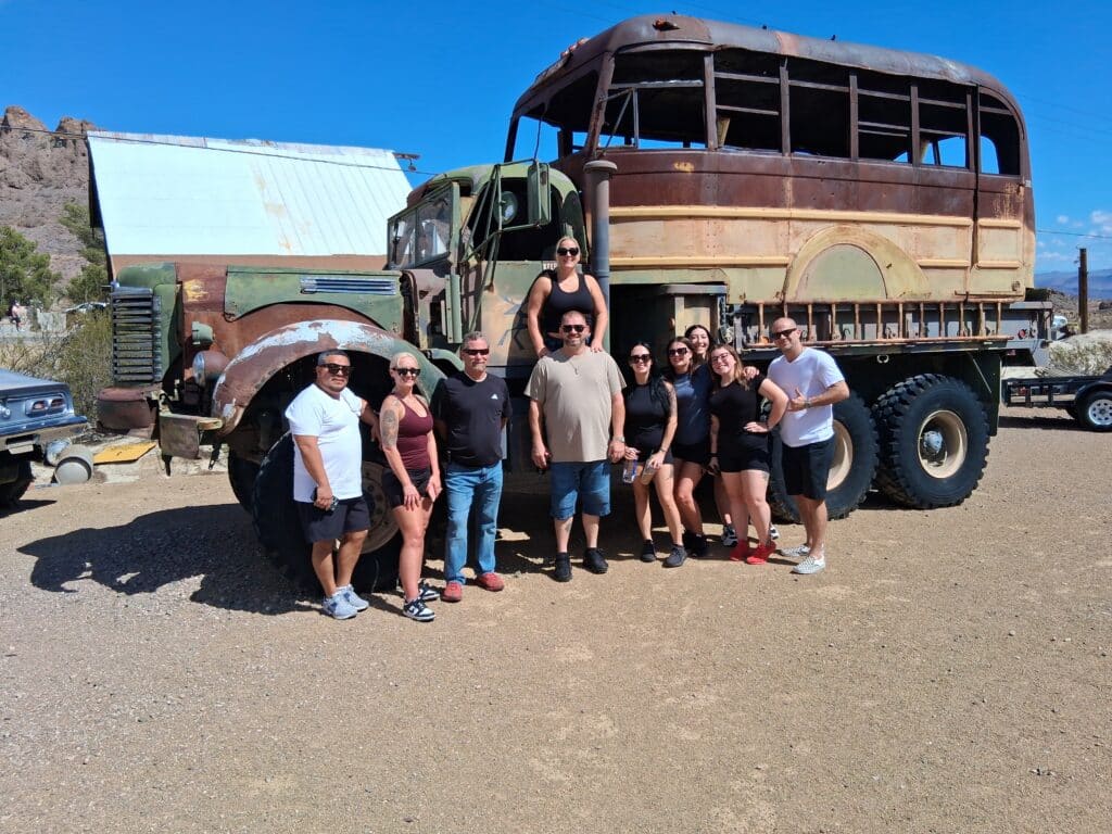 Ten people smiling by a rugged off-road vehicle in Nevada near a warehouse, ready for Las Vegas ATV and Ghost Town tours.