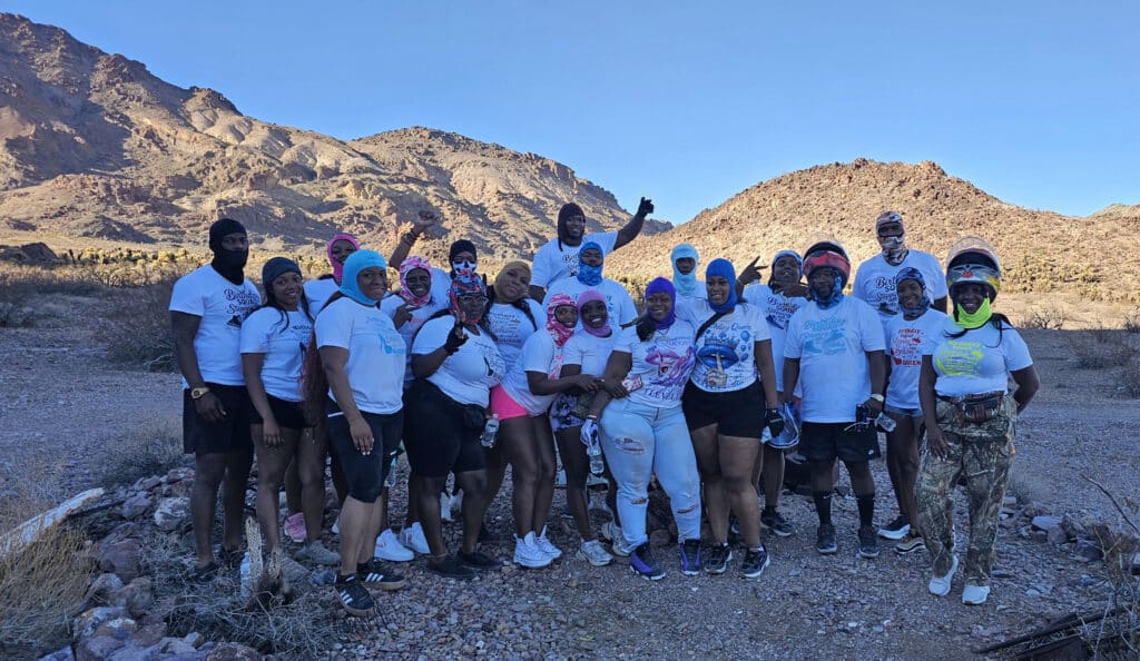 Group in white shirts poses on Nevada rocky terrain with mountains, blue sky; Colorado River ATV tour near Las Vegas.
