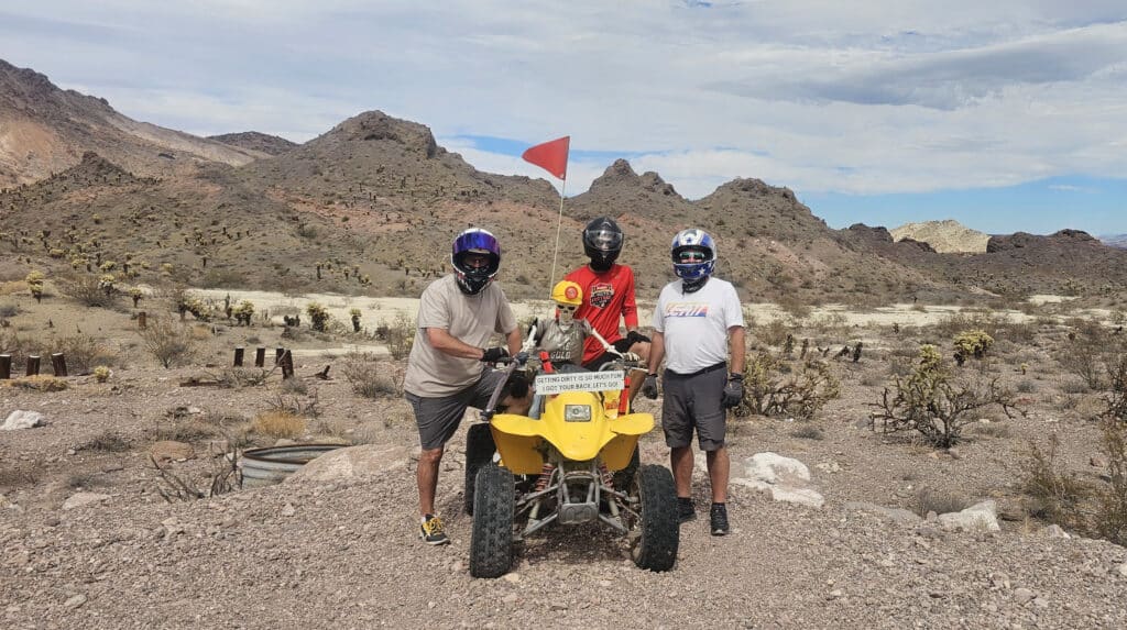 ATV riders with helmets pose by a yellow ATV in Nevada’s rocky desert near Las Vegas, mountains and sky in the background.