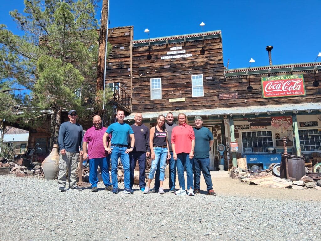 Nine men smile in front of a rustic Nevada ghost town with vintage signs, near Las Vegas, perfect for ATV tours or RZR rides.