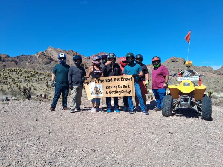 Eight people in helmets pose by a yellow ATV and sign in Nevada desert near Las Vegas, ready for off-road RZR adventure tours.