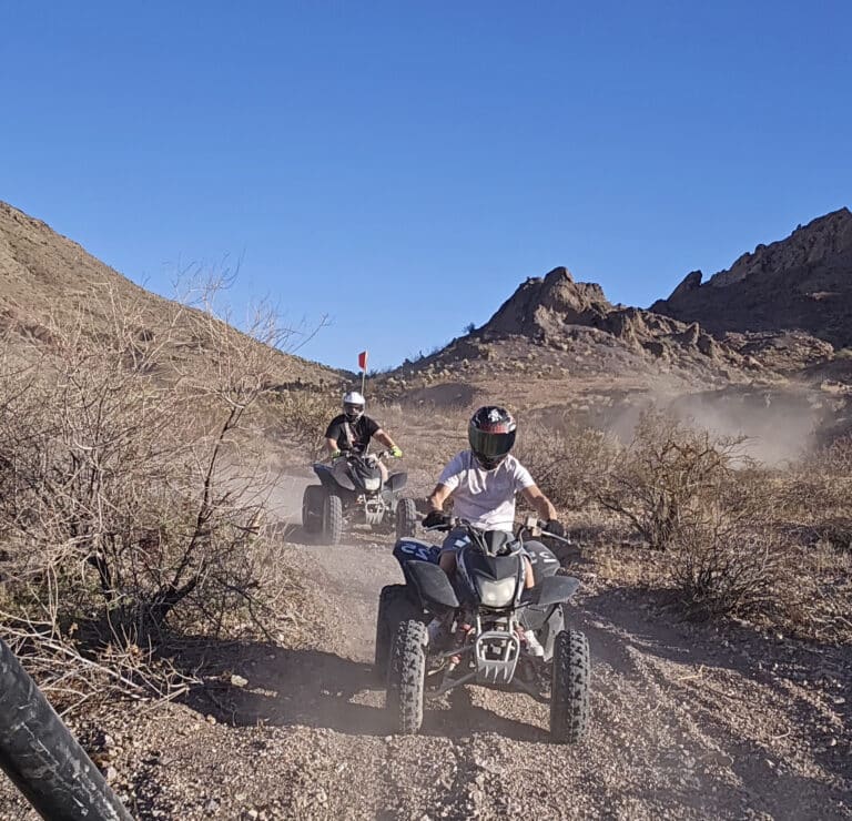 Adventurers on an ATV tour in Nevada ride dusty desert trails near Las Vegas, helmets on, rocky hills and dry bushes all around.