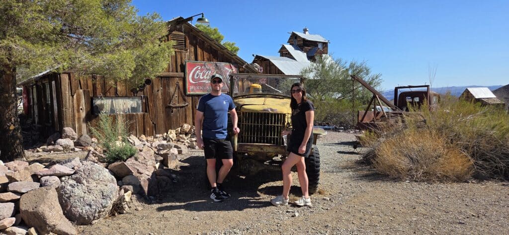 Two people in a Nevada ghost town stand by a rusty truck, vintage signs, and desert plants—perfect for Las Vegas sightseeing tours.