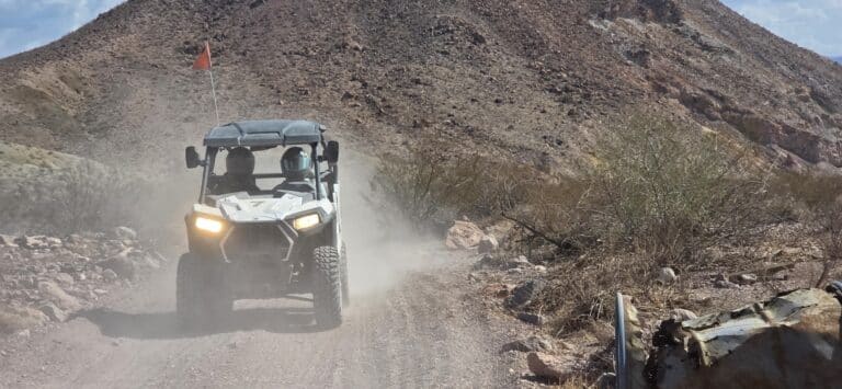 RZR off-road utility vehicle with two riders drives a dusty Nevada desert trail near Las Vegas, headlights on, dust swirling behind.