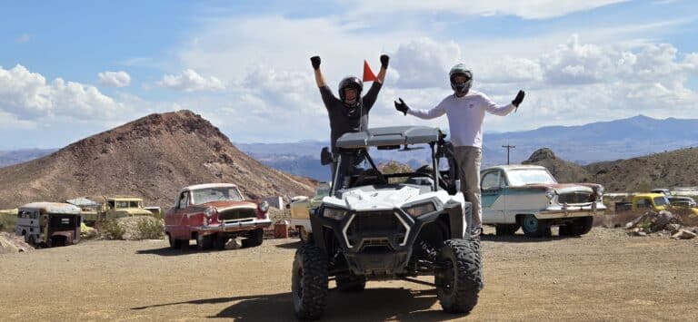 Adventurers in helmets pose on an ATV near Las Vegas, Nevada, with vintage cars, mountains, and desert skies—perfect for RZR tours.