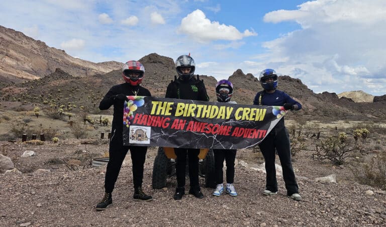 Four friends in racing suits with helmets hold a Birthday Crew banner on a rocky Nevada desert ATV tour near Las Vegas.