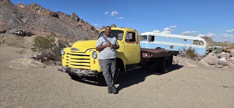 Smiling person by a vintage yellow truck, rocky Nevada desert near Las Vegas with ghost town sights and blue sky.