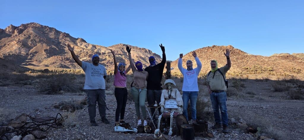 Group poses playfully in a Nevada desert near Las Vegas; skeleton with sign hints at ghost town sightseeing adventure.