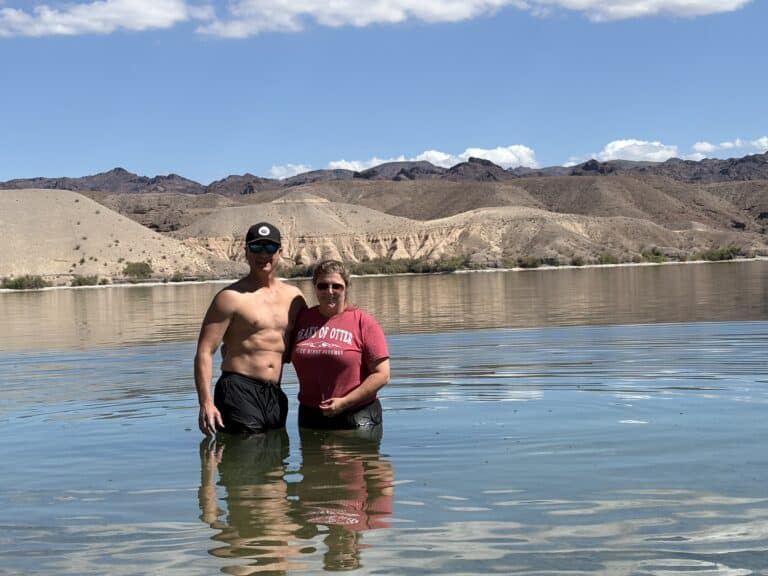 Couple smiles at camera in shallow Colorado River near Las Vegas, Nevada; rocky hills and cloudy sky in the background.