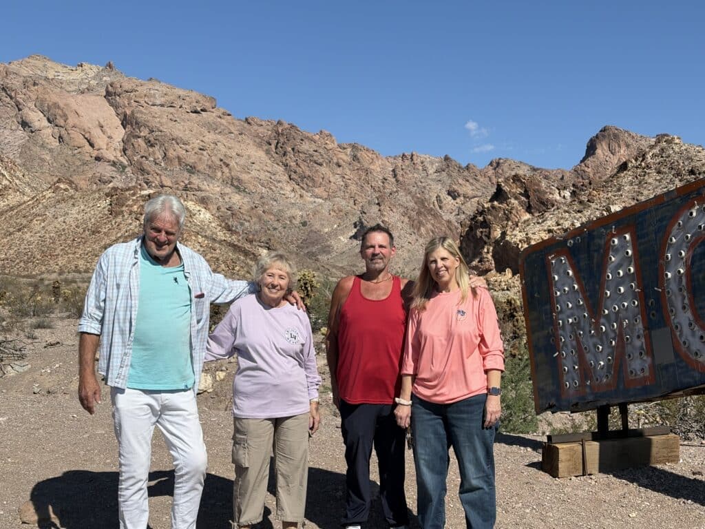Four smiling adults pose by rocky Nevada hills near Las Vegas, with old sign; perfect for Ghost Town sightseeing adventures.