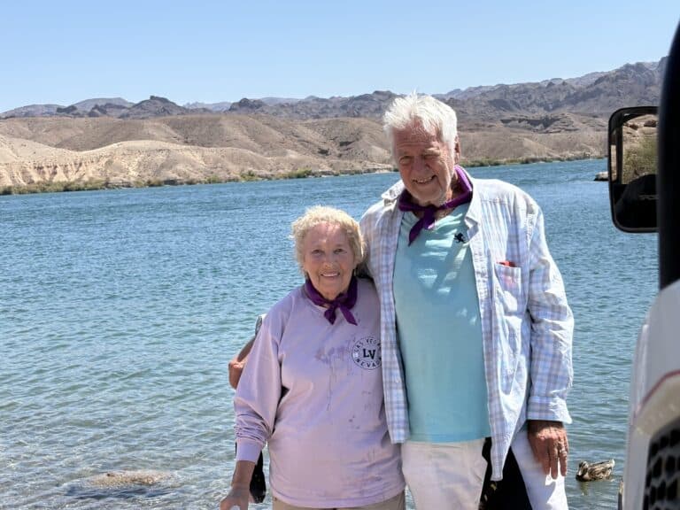 Smiling older couple in casual wear and purple bandanas by Nevada’s clear Colorado River, perfect for ATV tours near Las Vegas.