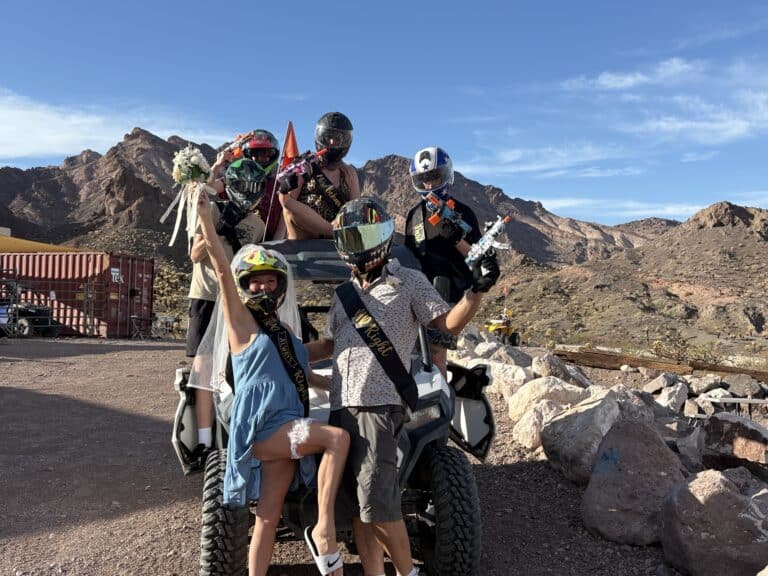 Group in helmets and costumes poses on a RZR during a Nevada ATV tour near Las Vegas, with desert mountains and blue skies.